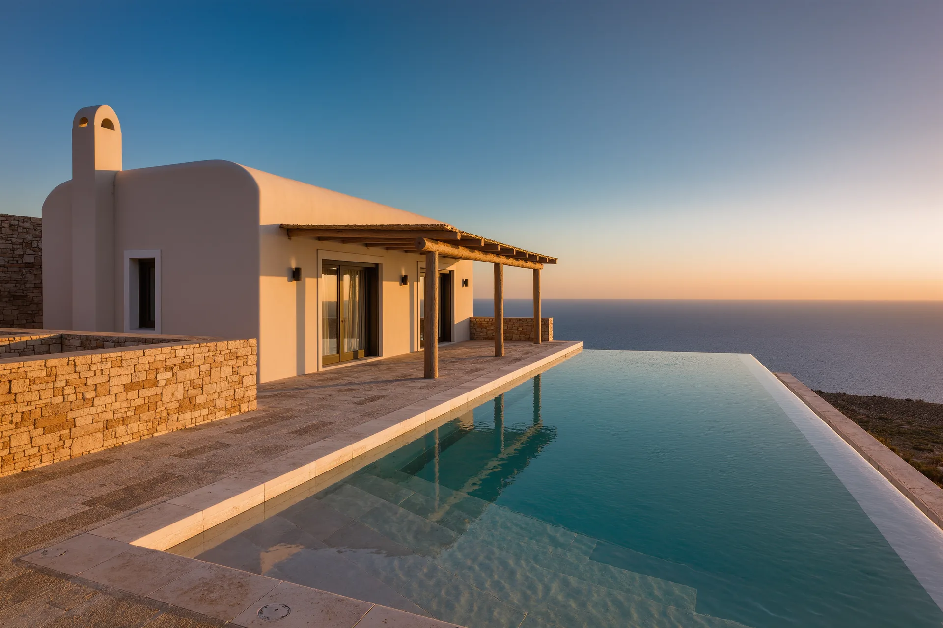 Villa aislada frente al mar al atardecer, sin otras construcciones a la vista, con una terraza de piedra y una piscina infinita que se funde con el horizonte