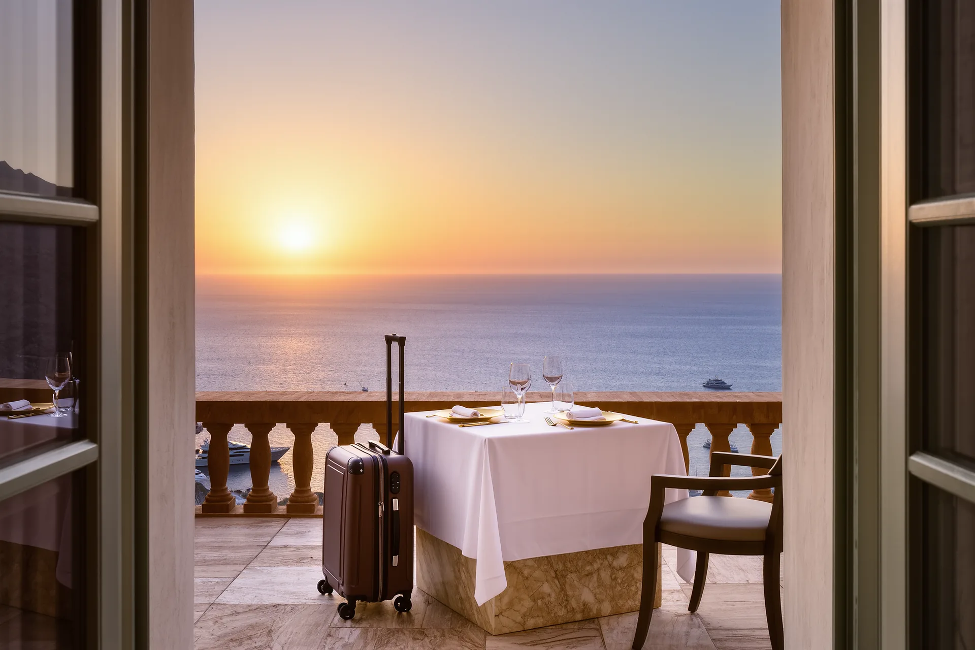 Terraza de un hotel frente al mar Mediterráneo al atardecer, con una mesa puesta para dos y maletas de cuero junto a la puerta